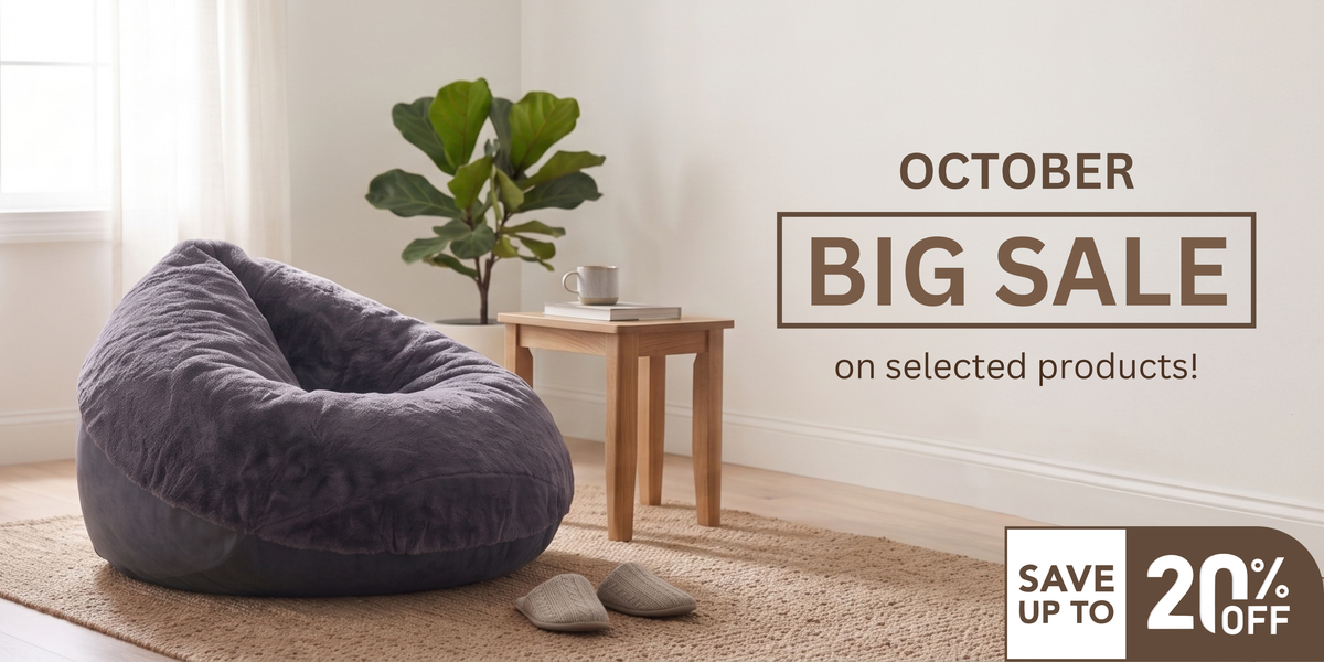 A dark gray plush bean bag chair sits on a beige rug in a bright room with a small wooden side table, a potted plant, and a pair of shoes nearby. Text on the right reads 'OCTOBER BIG SALE on selected products!' with a 'SAVE UP TO 20% OFF' badge.